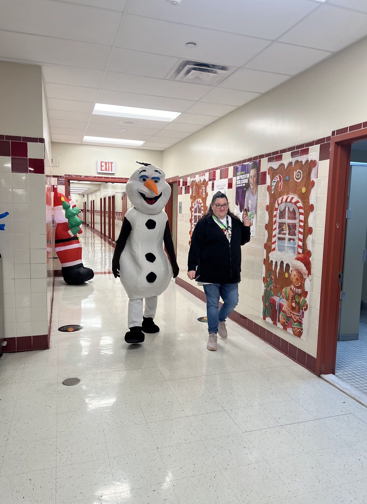A costumed snowman character walks down a decorated school hallway beside an adult.