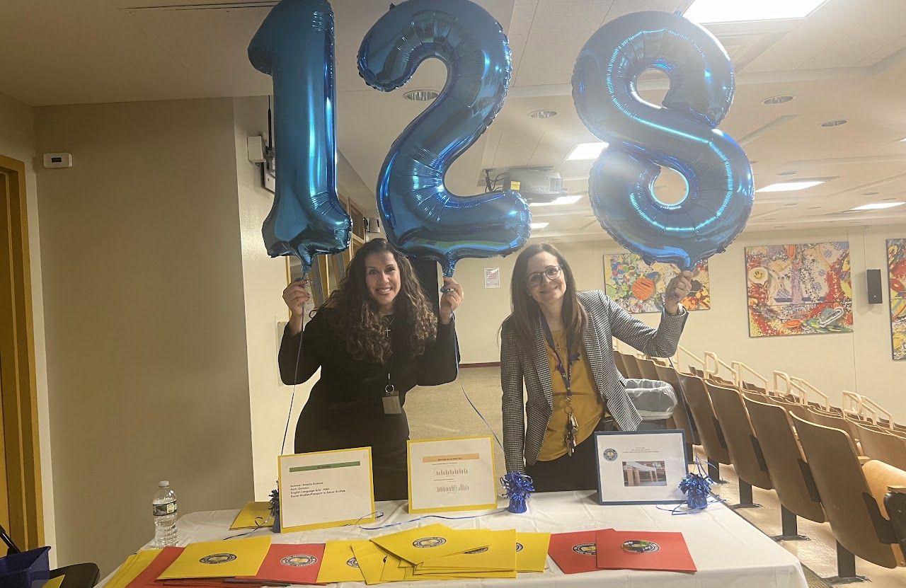 Two staff members smile behind a welcome table in the auditorium, holding blue 128 balloons.