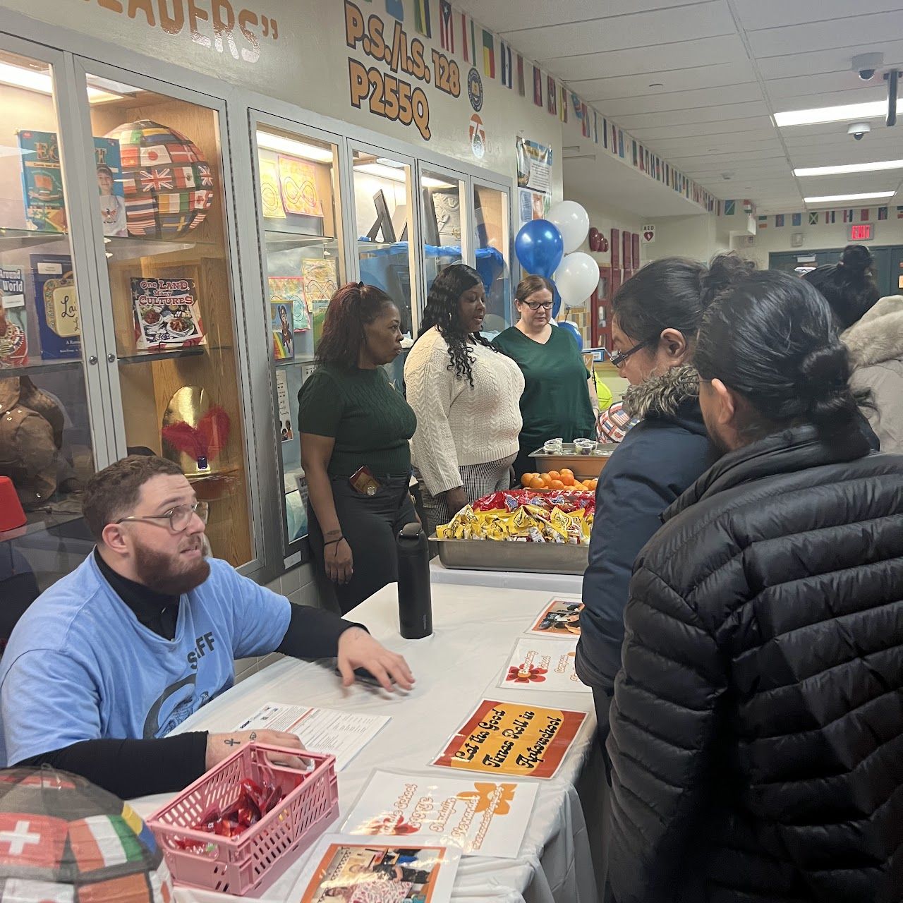 A staff member at a table of flyers and snacks talks with visiting families.